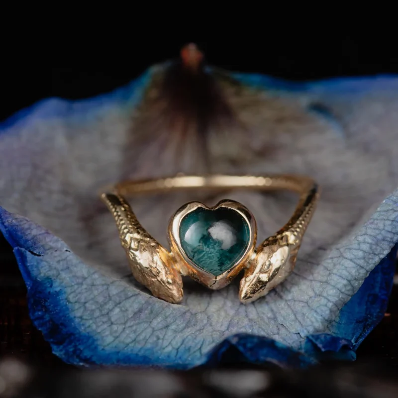 An image showing a 14K gold ring featuring a heart shaped london blue topaz and a snake in each side of the ring, photographed inside a petal of a blue rose