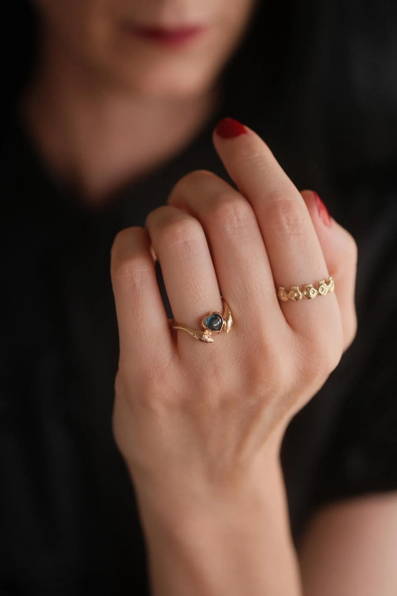 An image showing a 14K gold ring featuring a heart shaped london blue topaz and a snake in each side of the ring from the side on a finger