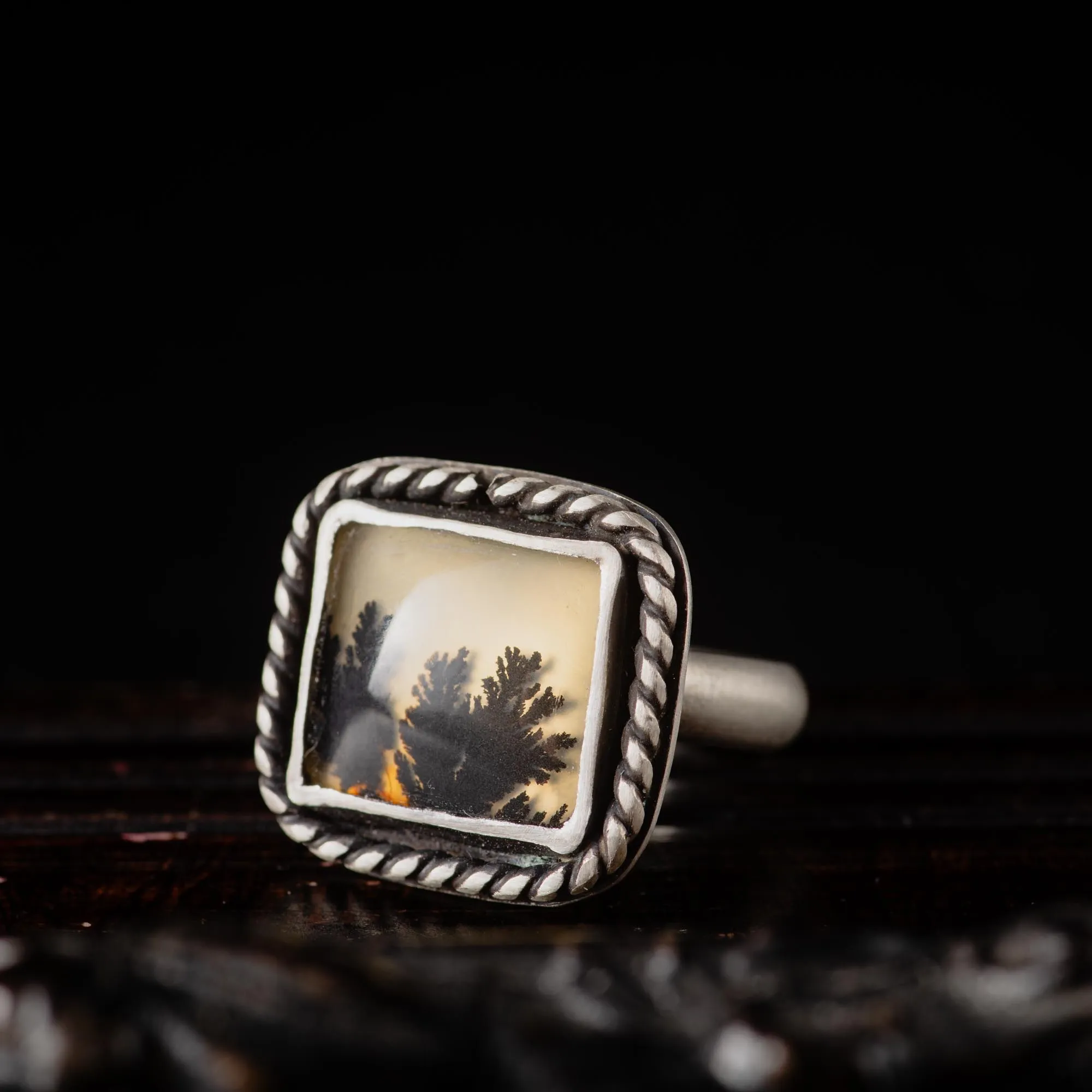 An image showing a rectangular dendritic agate gemstone with twisted border on a sterling silver ring from the side