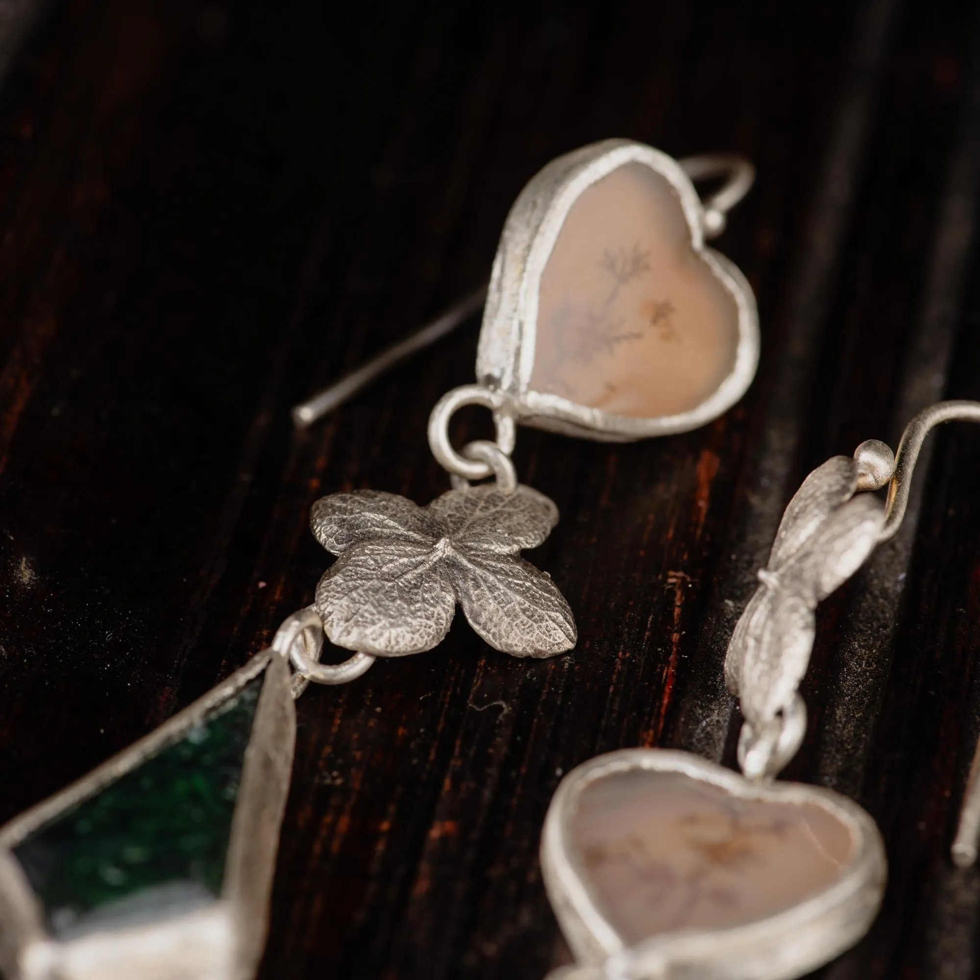 An image showing details of a sterling silver pair of earrings featuring dendritic agate in heart shaped and moss agate in kite shaped