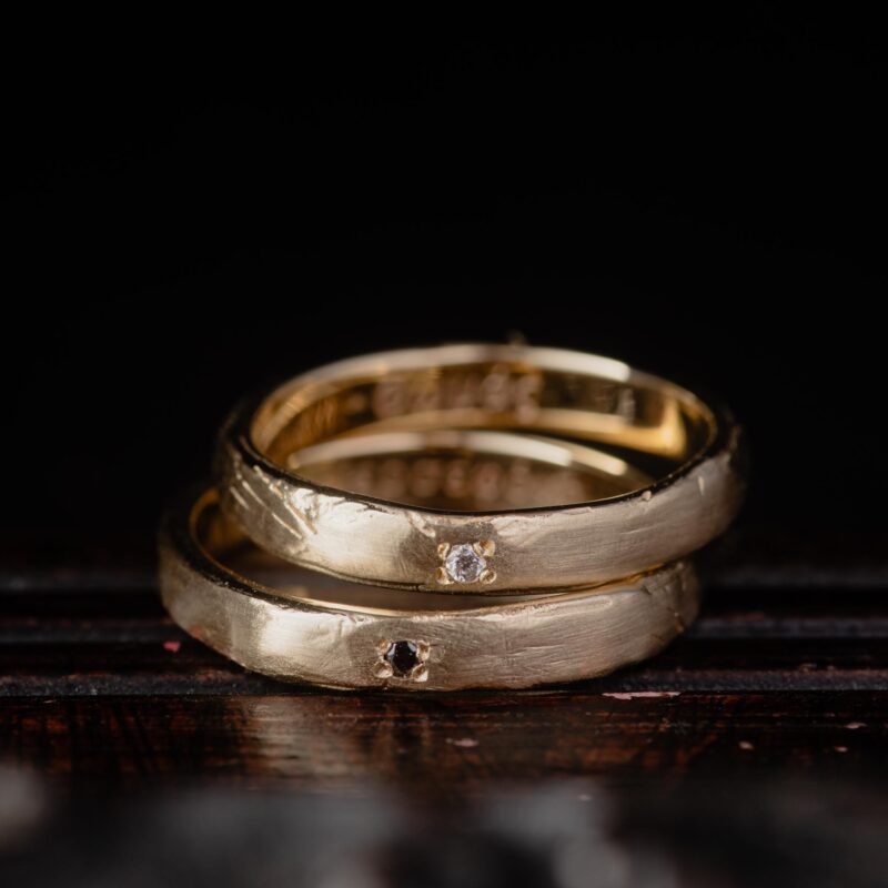 An image showing two wedding bands in organic form in 14K yellow gold , one with white diamond, the other with black diamond
